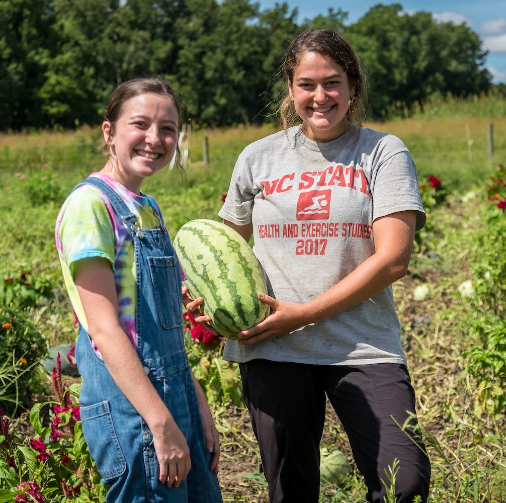 two NC State students hold a watermelon while standing in a garden at the Field Lab